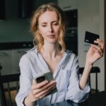 A woman holds a credit card while looking at her smartphone at home.