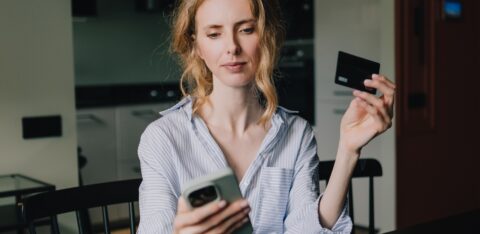 A woman holds a credit card while looking at her smartphone at home.