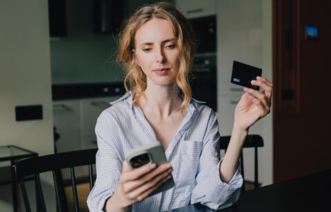 A woman holds a credit card while looking at her smartphone at home.
