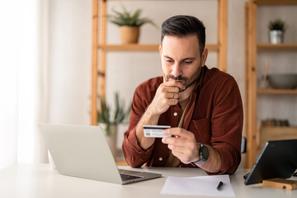 Serious business man sitting at table in home office, holding and looking at credit card, worried about debt and credit card deficit, considering a financial loan from bank.
