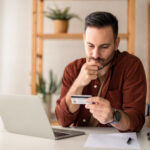 Serious business man sitting at table in home office, holding and looking at credit card, worried about debt and credit card deficit, considering a financial loan from bank.