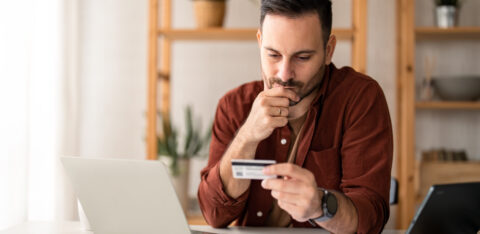 Serious business man sitting at table in home office, holding and looking at credit card, worried about debt and credit card deficit, considering a financial loan from bank.