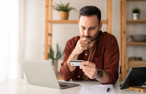 Serious business man sitting at table in home office, holding and looking at credit card, worried about debt and credit card deficit, considering a financial loan from bank.