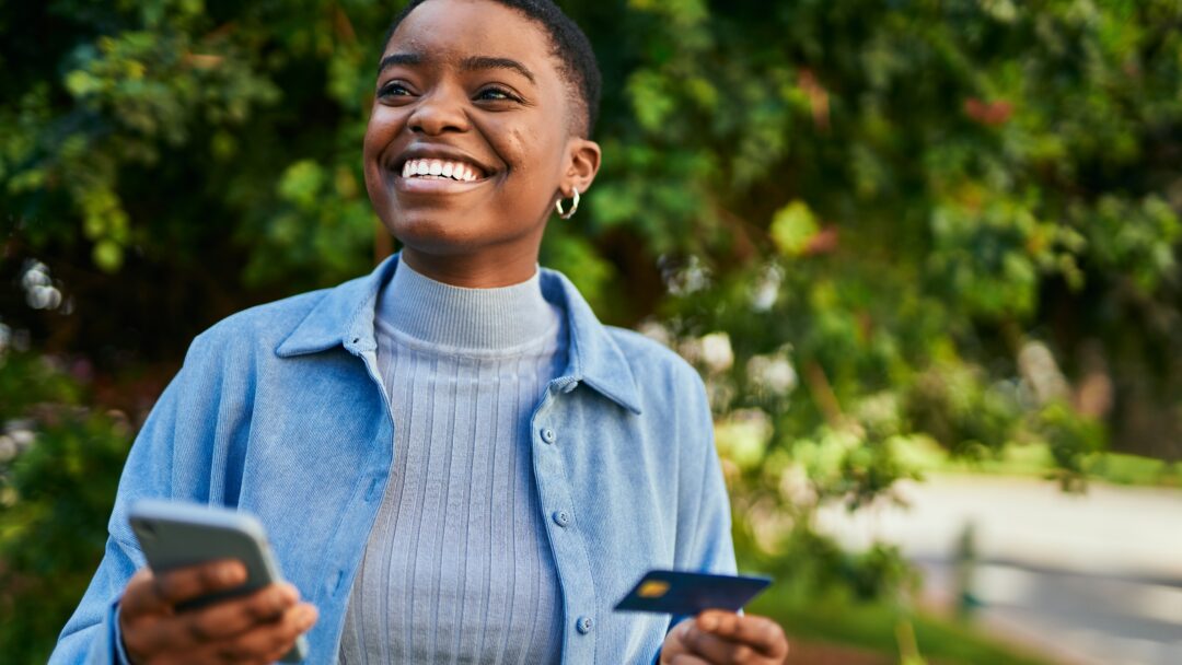 Young african american woman smiling happy holding smartphone and credit card at the city