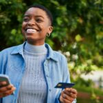 Young african american woman smiling happy holding smartphone and credit card at the city