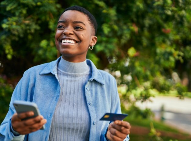 Young african american woman smiling happy holding smartphone and credit card at the city