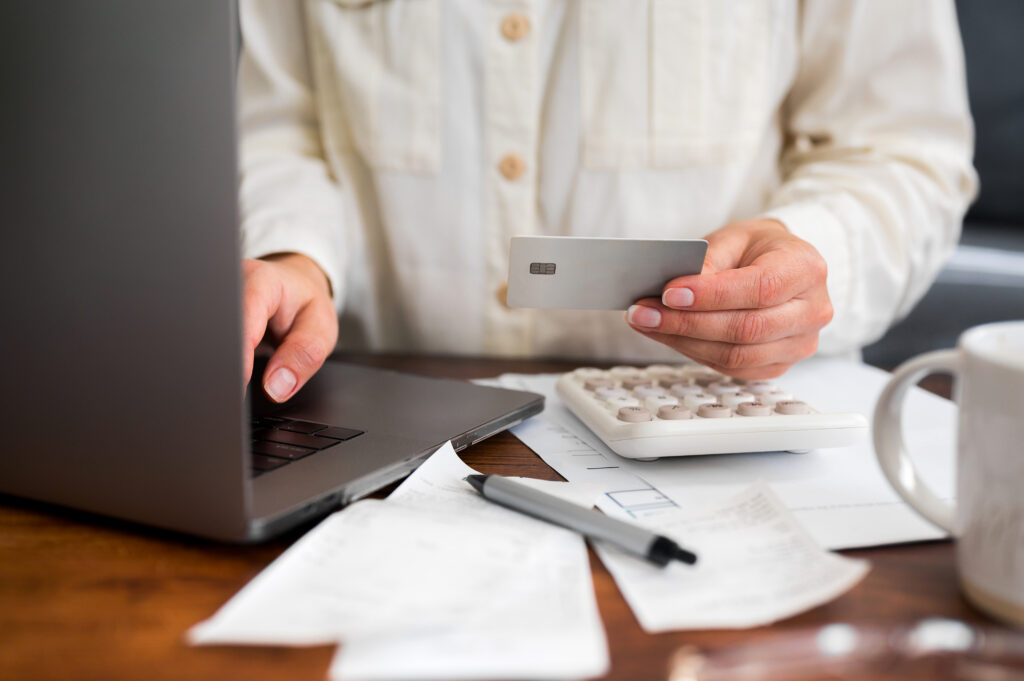 Person holding a credit card while using a laptop, with a calculator and bills on the desk.