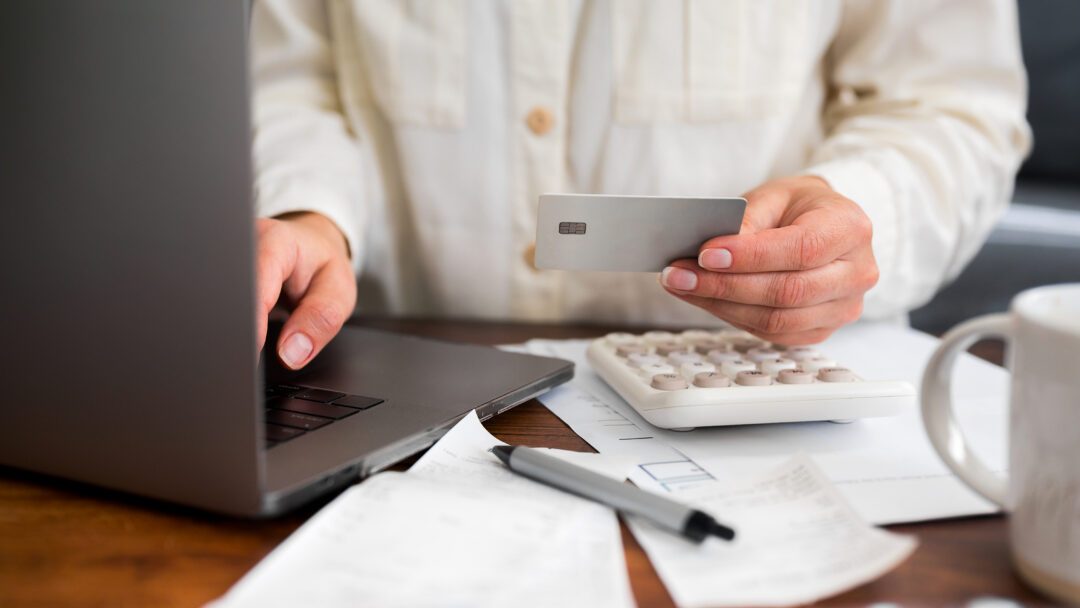 Person holding a credit card while using a laptop, with a calculator and bills on the desk.