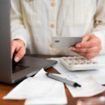 Person holding a credit card while using a laptop, with a calculator and bills on the desk.