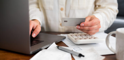 Person holding a credit card while using a laptop, with a calculator and bills on the desk.
