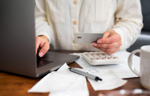 Person holding a credit card while using a laptop, with a calculator and bills on the desk.