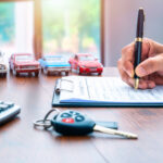 Man signing contract on car loan application form. Car loan or buying a new car concept. Miniature cars and car key on table.