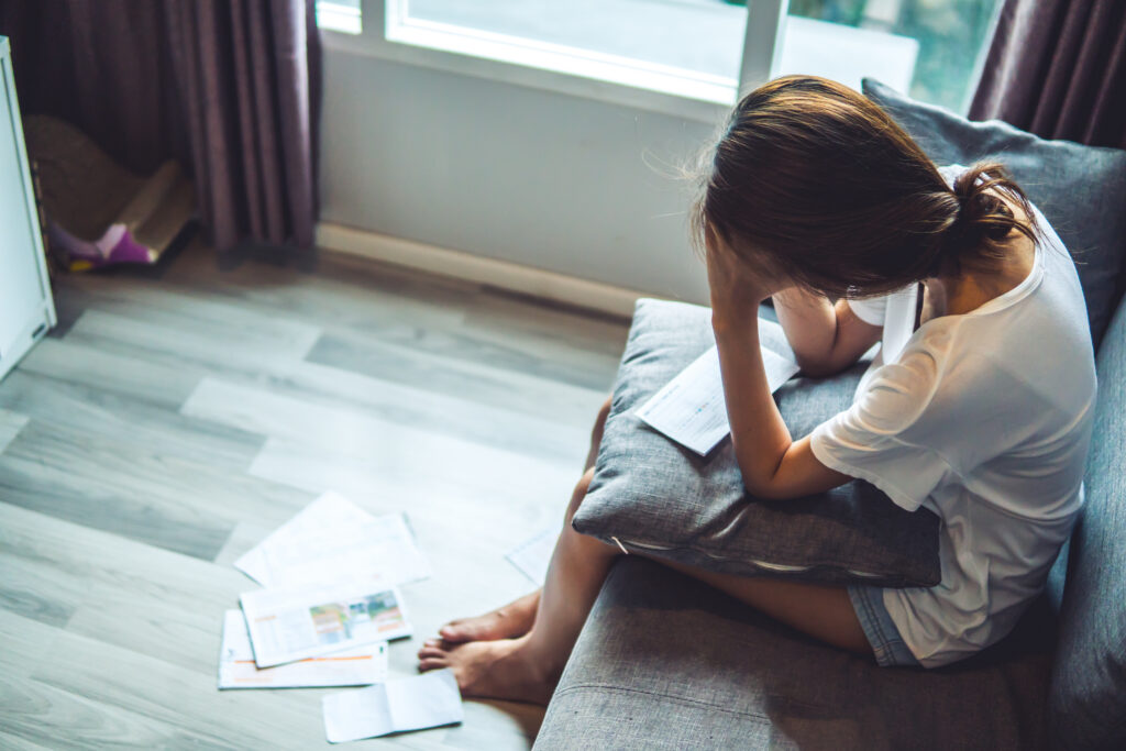 Stressed woman sitting on a couch with bills and paperwork scattered nearby.