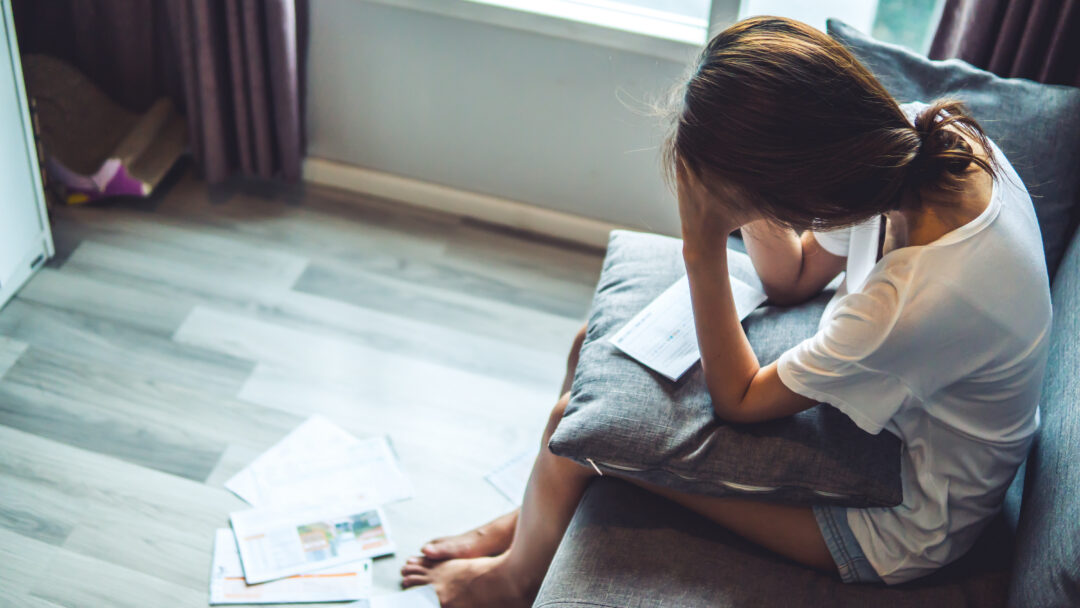 Stressed woman sitting on a couch with bills and paperwork scattered nearby.