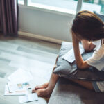 Stressed woman sitting on a couch with bills and paperwork scattered nearby.