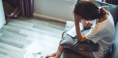 Stressed woman sitting on a couch with bills and paperwork scattered nearby.