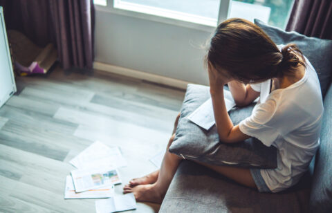 Stressed woman sitting on a couch with bills and paperwork scattered nearby.