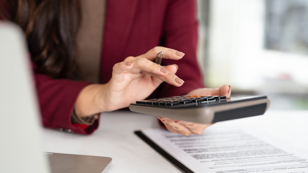 Woman in a burgundy blazer using a calculator while reviewing financial documents at her desk.
