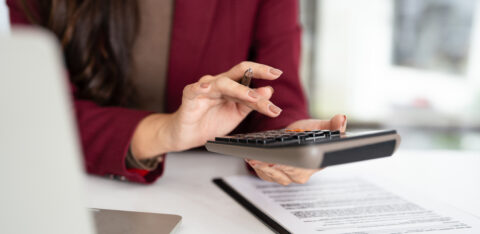Woman in a burgundy blazer using a calculator while reviewing financial documents at her desk.