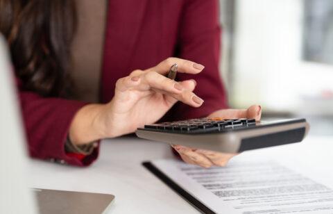 Woman in a burgundy blazer using a calculator while reviewing financial documents at her desk.