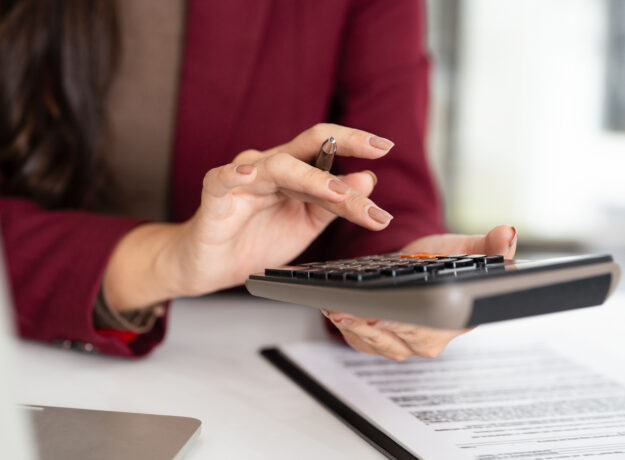 Woman in a burgundy blazer using a calculator while reviewing financial documents at her desk.
