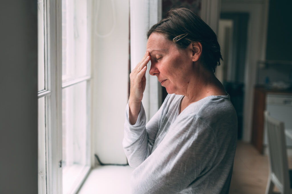 Stressed woman standing by a window with her hand on her forehead.