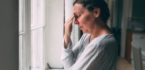 Stressed woman standing by a window with her hand on her forehead.