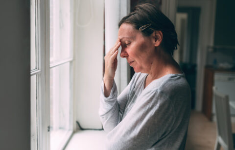 Stressed woman standing by a window with her hand on her forehead.