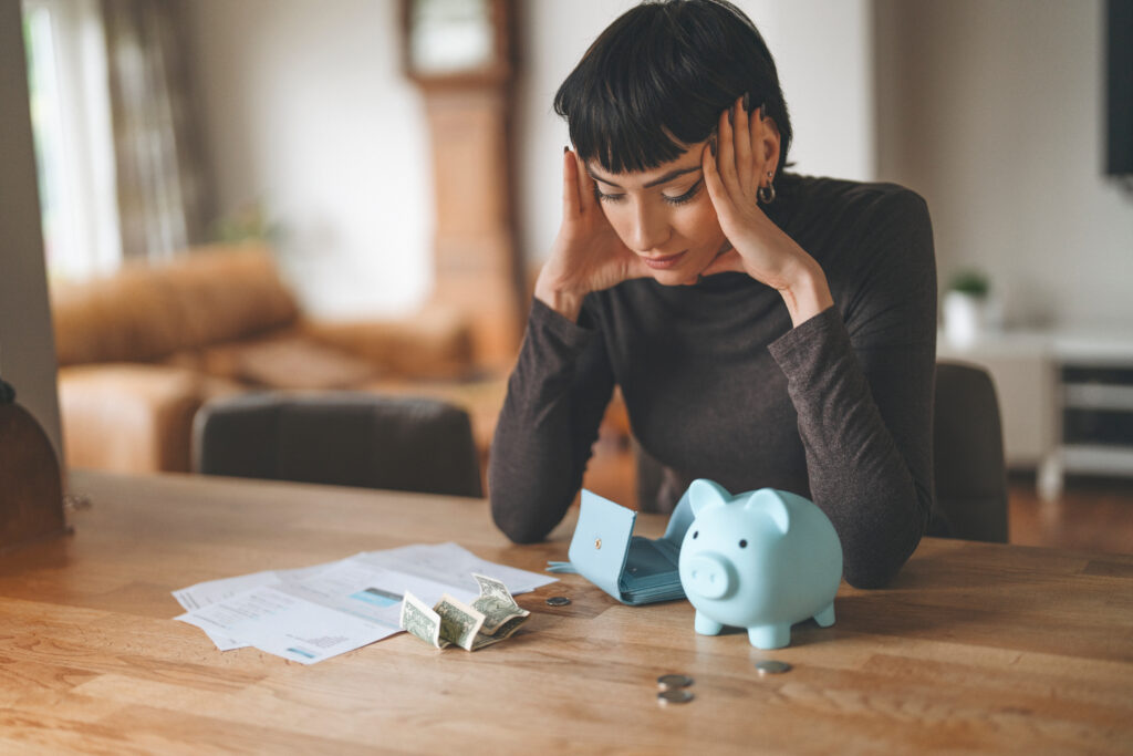 Woman sitting at a table with bills, cash, an empty wallet, and a piggy bank while reviewing her finances.