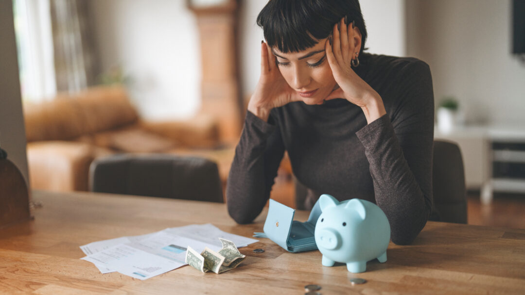 Woman sitting at a table with bills, cash, an empty wallet, and a piggy bank while reviewing her finances.