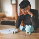 Woman sitting at a table with bills, cash, an empty wallet, and a piggy bank while reviewing her finances.