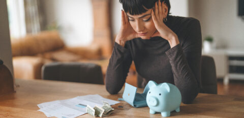Woman sitting at a table with bills, cash, an empty wallet, and a piggy bank while reviewing her finances.