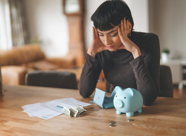 Woman sitting at a table with bills, cash, an empty wallet, and a piggy bank while reviewing her finances.