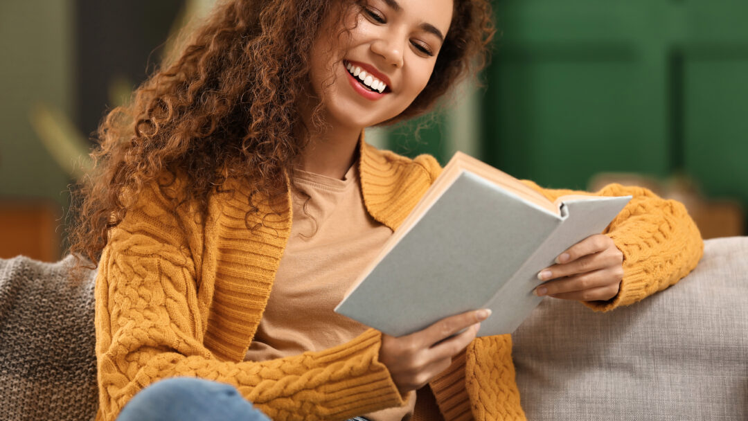 Young African American woman in yellow cardigan reading book on sofa at home