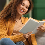 Young African American woman in yellow cardigan reading book on sofa at home