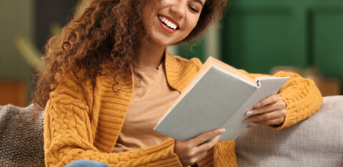 Young African American woman in yellow cardigan reading book on sofa at home