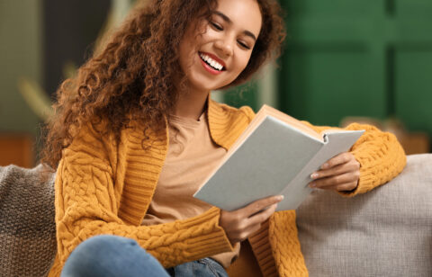 Young African American woman in yellow cardigan reading book on sofa at home