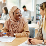 Happy Muslim student talking to her classmate both sitting at a desk