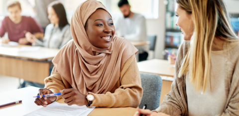 Happy Muslim student talking to her classmate both sitting at a desk