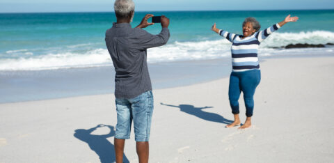 Front view of an African American couple standing on the beach with blue sky and sea in the background, the woman raising her hands and smiling, while the man takes a photo of her with his smartphone