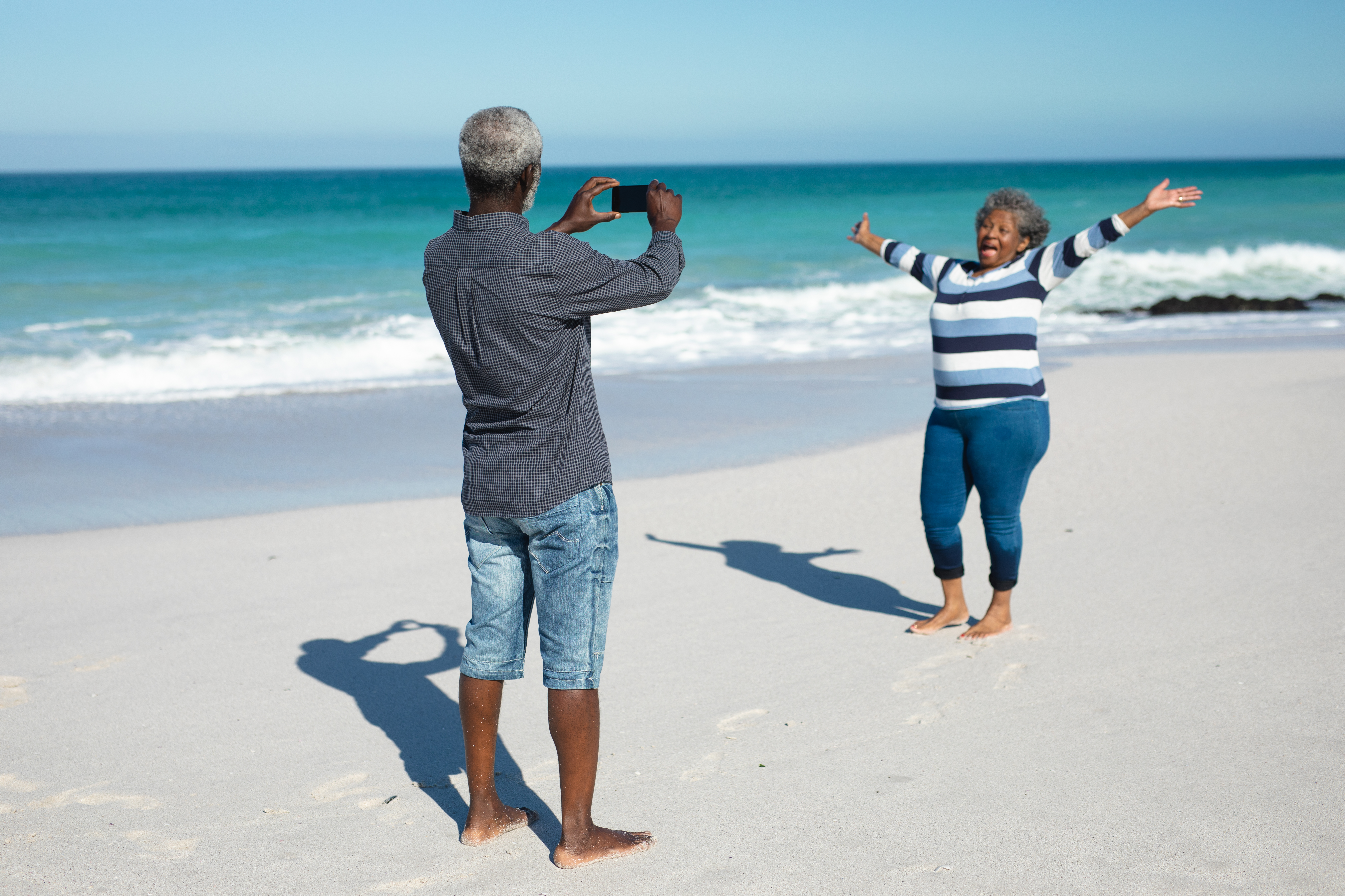 Front view of an African American couple standing on the beach with blue sky and sea in the background, the woman raising her hands and smiling, while the man takes a photo of her with his smartphone