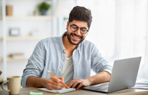 Happy arab man writing business report on laptop and taking notes, sitting at workplace at home interior
