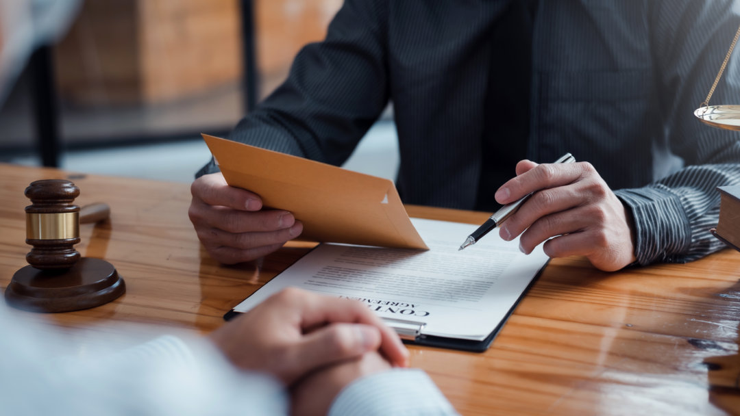 Lawyer reviewing a legal document with a client, holding an envelope and a pen at a wooden desk.