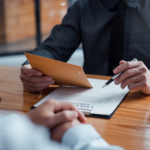 Lawyer reviewing a legal document with a client, holding an envelope and a pen at a wooden desk.