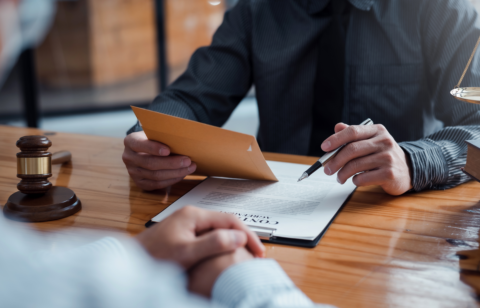Lawyer reviewing a legal document with a client, holding an envelope and a pen at a wooden desk.