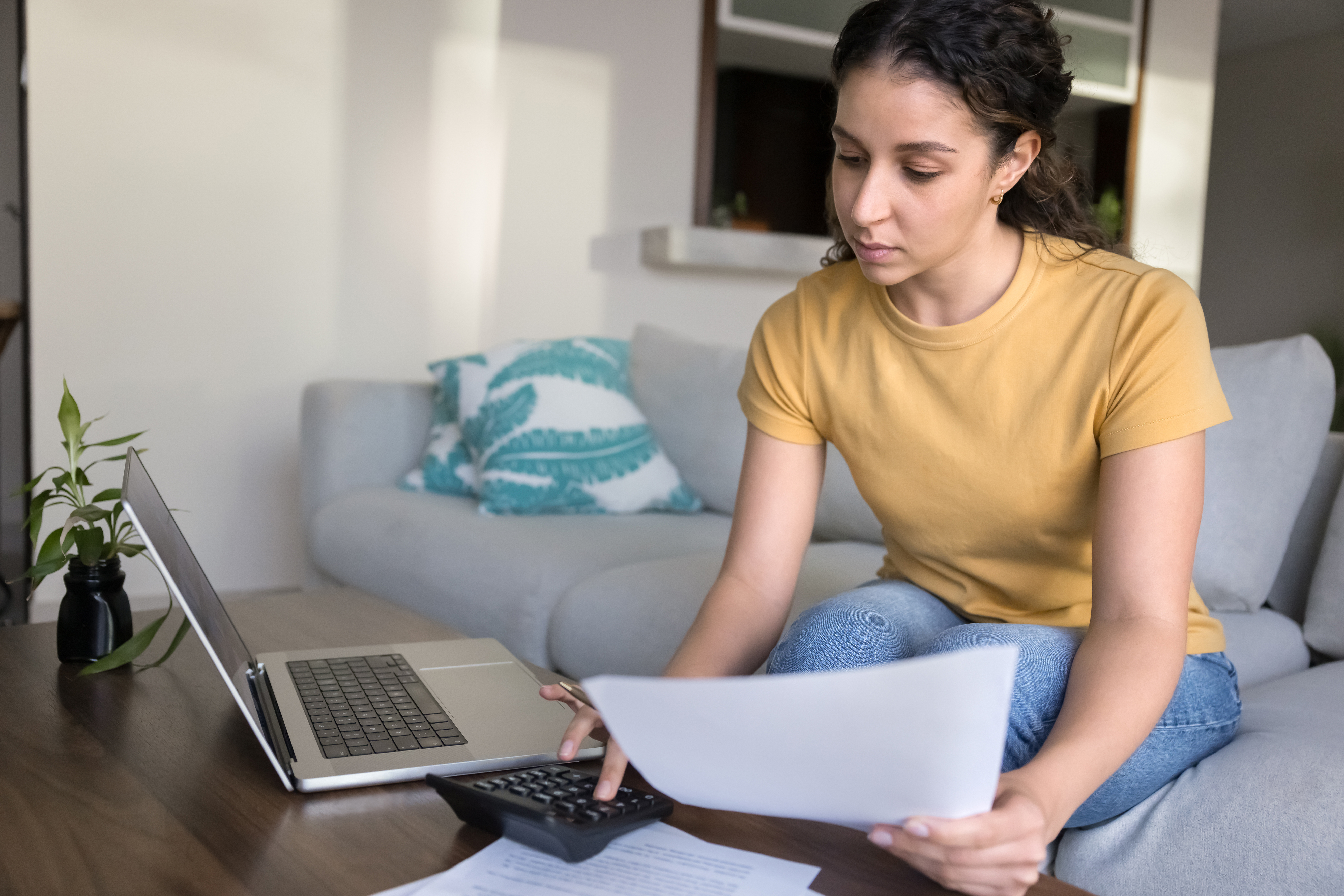 Young woman sitting on a couch reviewing financial documents with a calculator and laptop on the table.