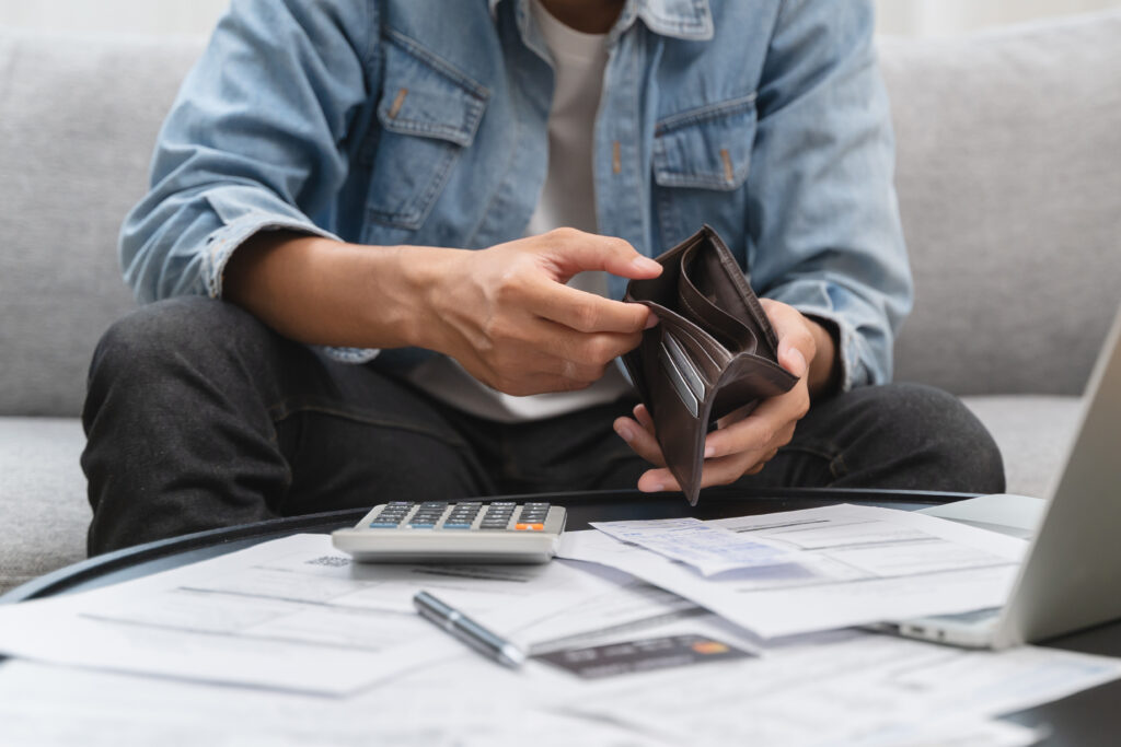 Man sitting on a couch checking his empty wallet, surrounded by bills, a calculator, and a laptop.