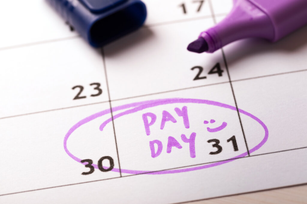 Close-up of a calendar with "PAY DAY" written in purple marker on the 31st, circled with a smiley face, alongside an open marker.