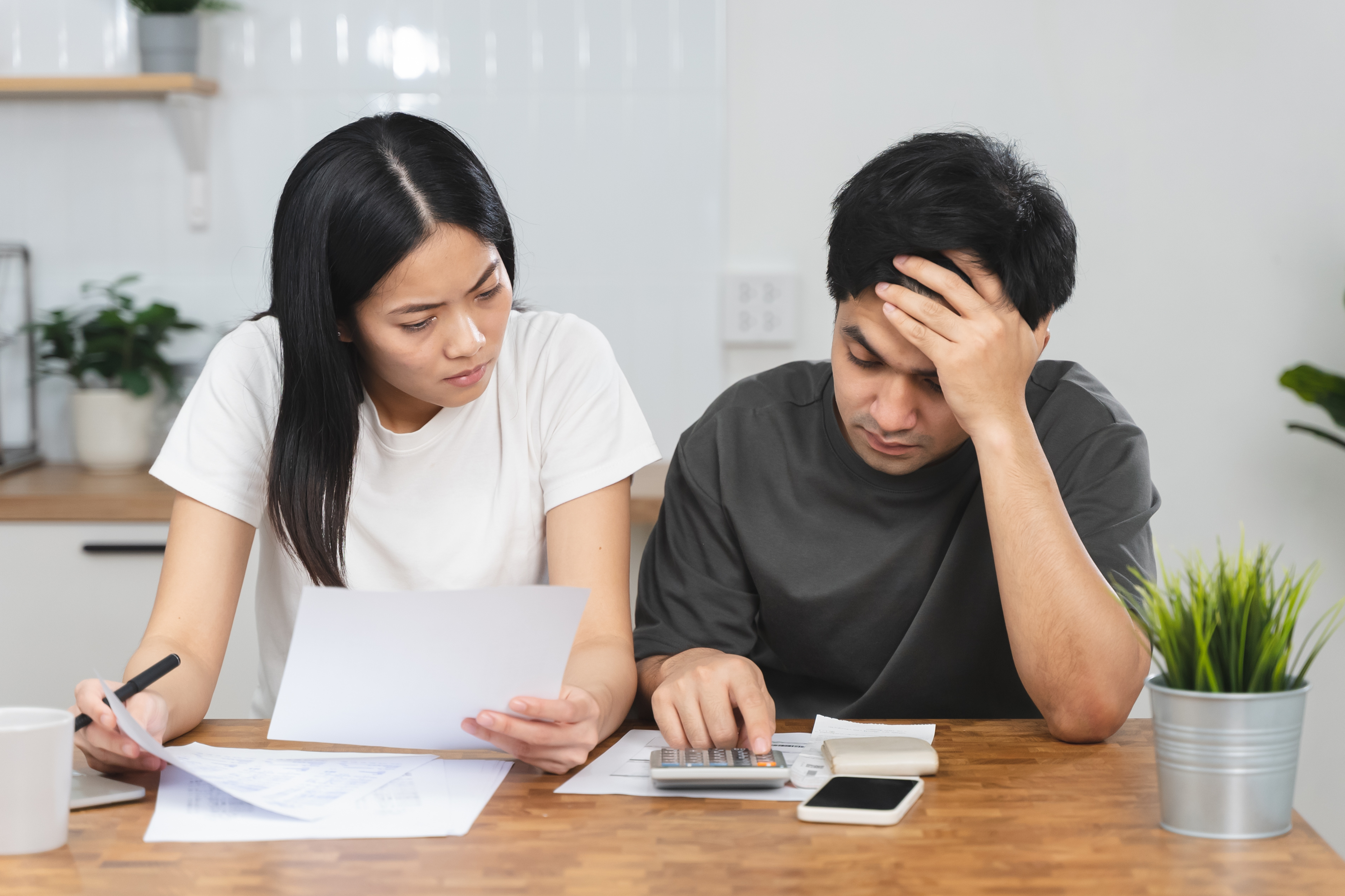 Stressed couple sitting at a table reviewing financial documents and using a calculator to manage expenses.