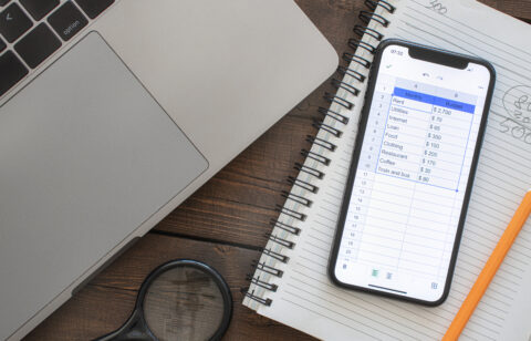 A smartphone displaying a digital budgeting spreadsheet next to a laptop, notebook, and pencil on a wooden desk.